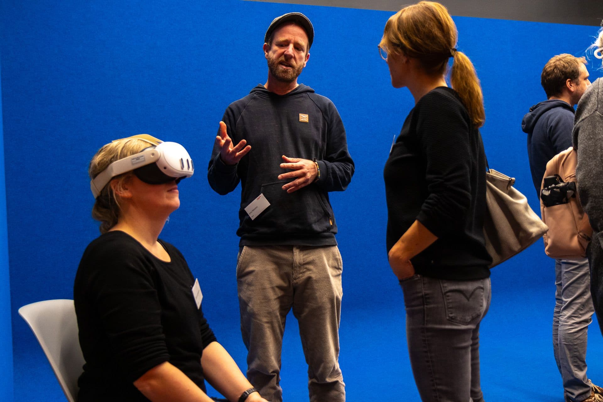 IWM-Alumni, die sich unterhalten, vor blauem Hintergrund. Person mit VR-Brille im Vordergrund.