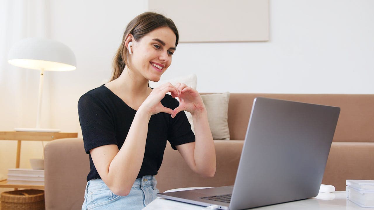 Eine Frau formt ein Herz mit ihren Händen und zeigt es in die Laptopkamera
