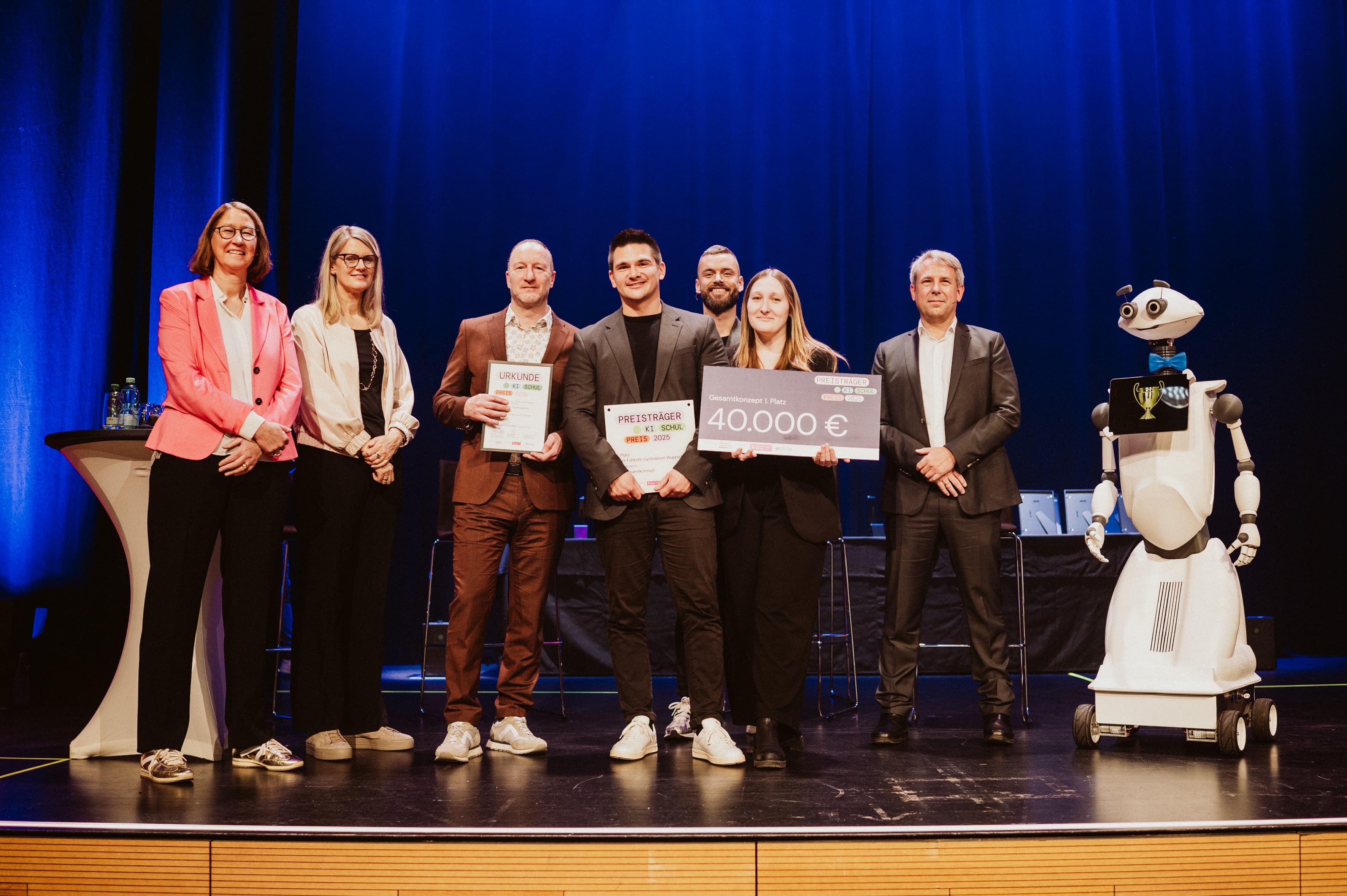“Award ceremony on stage: A group of eight people pose with certificates and a symbolic cheque worth €40,000. A humanoid robot stands on the right. The scene is set against blue stage lighting, documenting the award given to a project team.